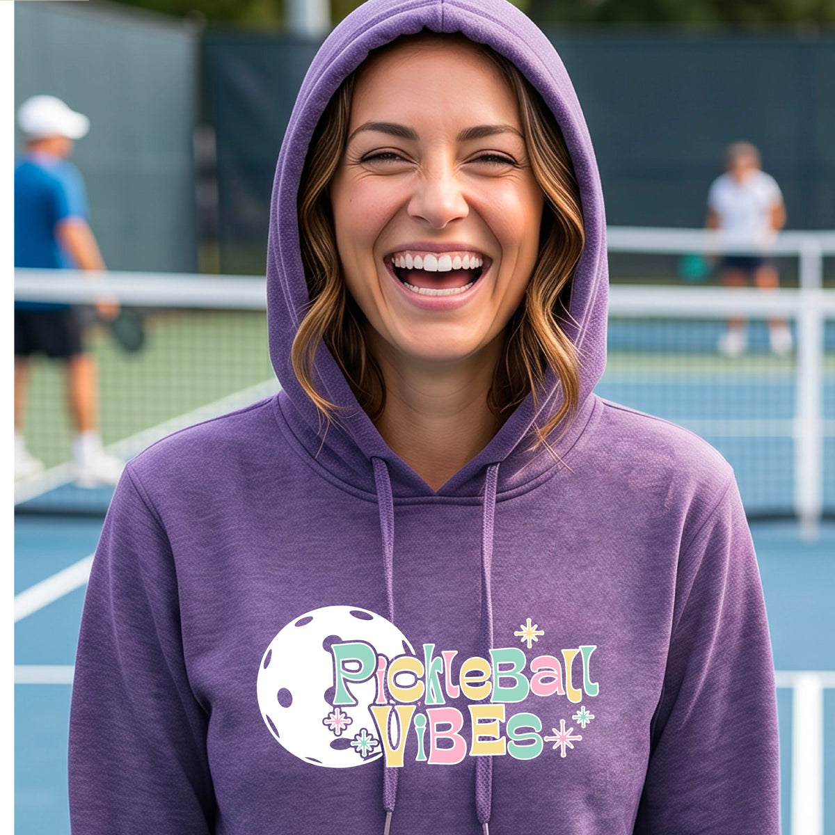 A close-up of a woman on a pickleball court wearing a purple fitted hoodie pullover, with a colorful retro style font 'Pickleball Vibes' on top of a white pickleball graphic, on the front of the hoodie. 