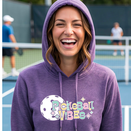 A close-up of a woman on a pickleball court wearing a purple fitted hoodie pullover, with a colorful retro style font 'Pickleball Vibes' on top of a white pickleball graphic, on the front of the hoodie. 