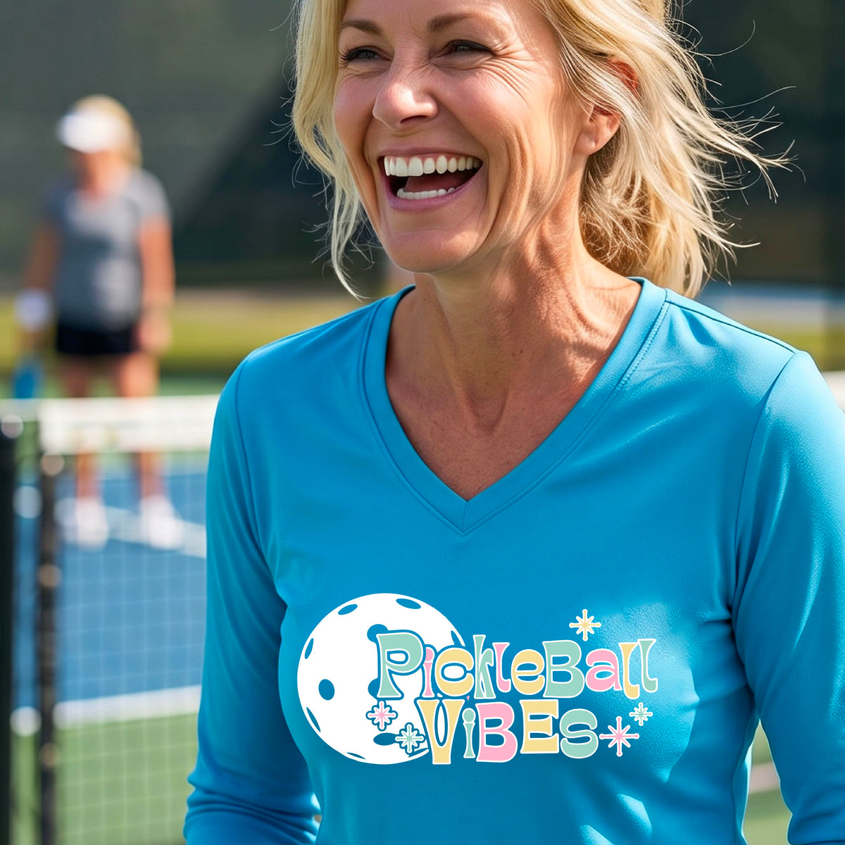 A close-up of a woman on a pickleball wearing a bright blue long-sleeve t-shirt, with a colorful retro style font 'Pickleball Vibes' on top of a white pickleball graphic, on the front of the shirt.