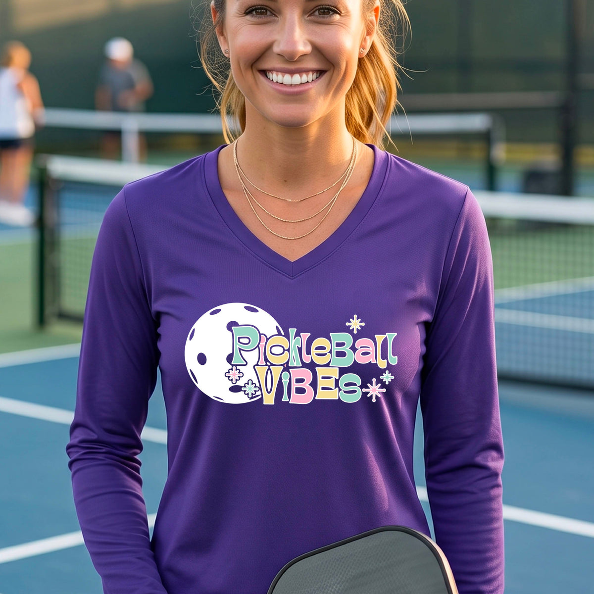 A close-up of a woman on a pickleball wearing a purple long-sleeve t-shirt, with a colorful retro style font 'Pickleball Vibes' on top of a white pickleball graphic, on the front of the shirt.