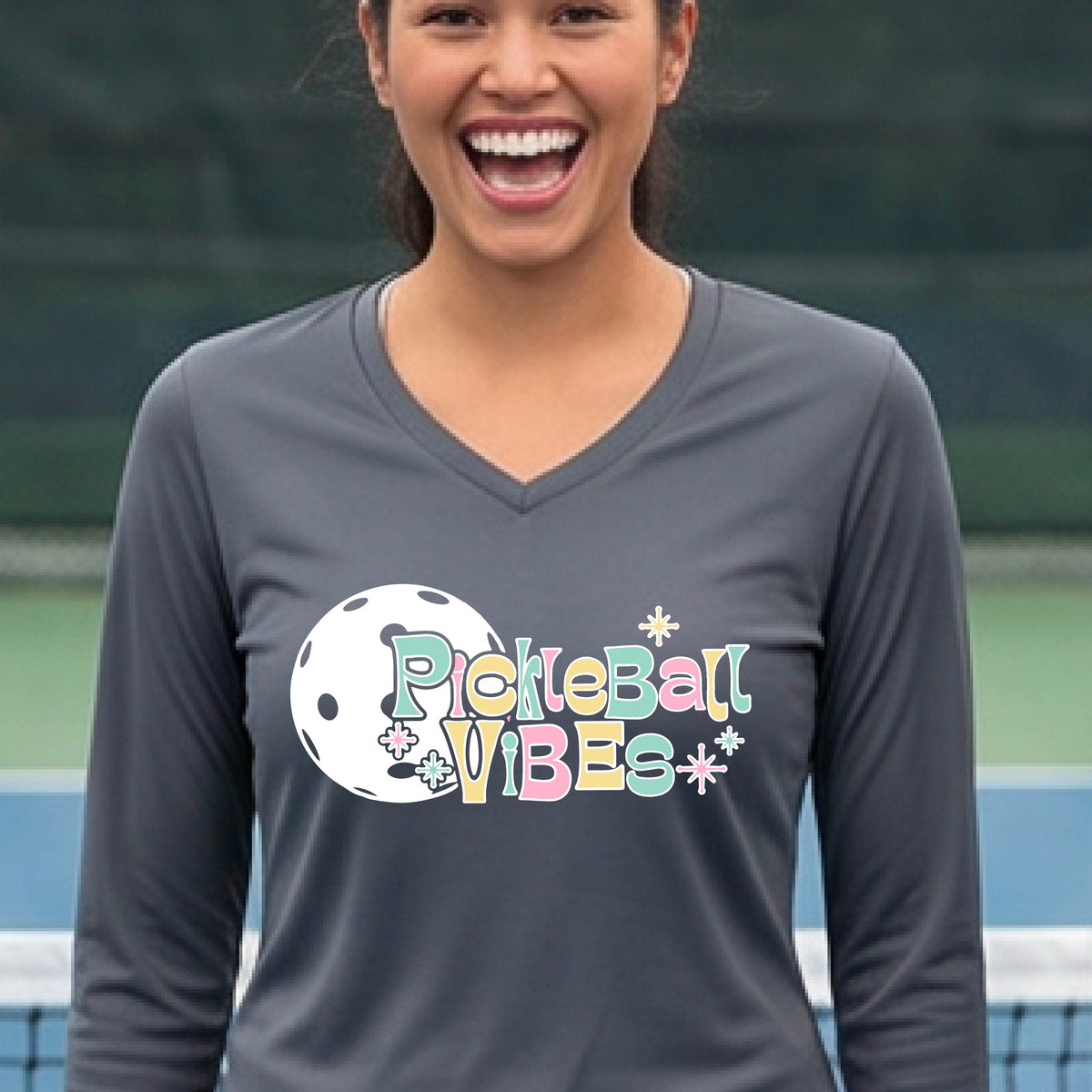 A close-up of a woman on a pickleball wearing a dark gray long-sleeve t-shirt, with a colorful retro style font 'Pickleball Vibes' on top of a white pickleball graphic, on the front of the shirt.