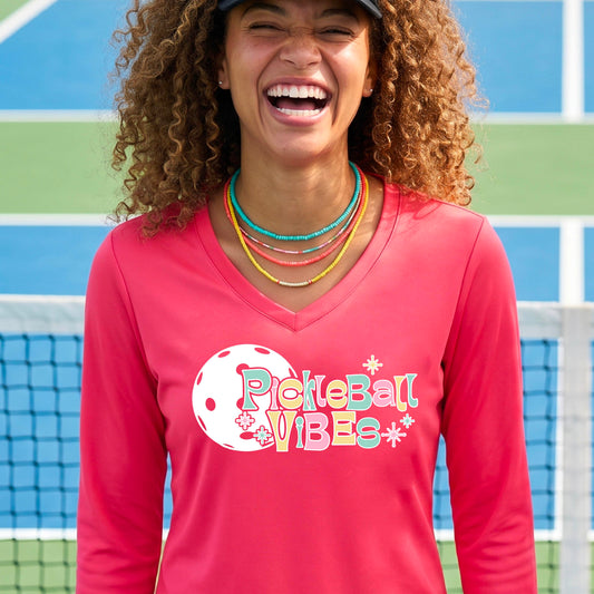 A close-up of a woman on a pickleball wearing a coral pink long-sleeve t-shirt, with a colorful retro style font 'Pickleball Vibes' on top of a white pickleball graphic, on the front of the shirt.