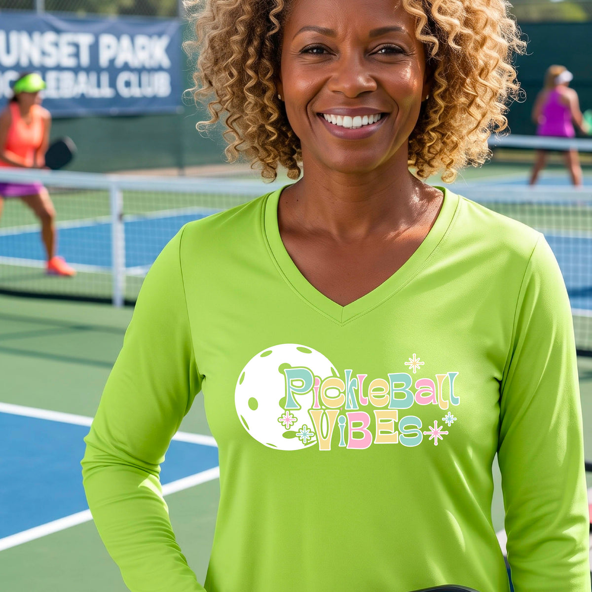 A close-up of a woman on a pickleball wearing a lime green long-sleeve t-shirt, with a colorful retro style font 'Pickleball Vibes' on top of a white pickleball graphic, on the front of the shirt.