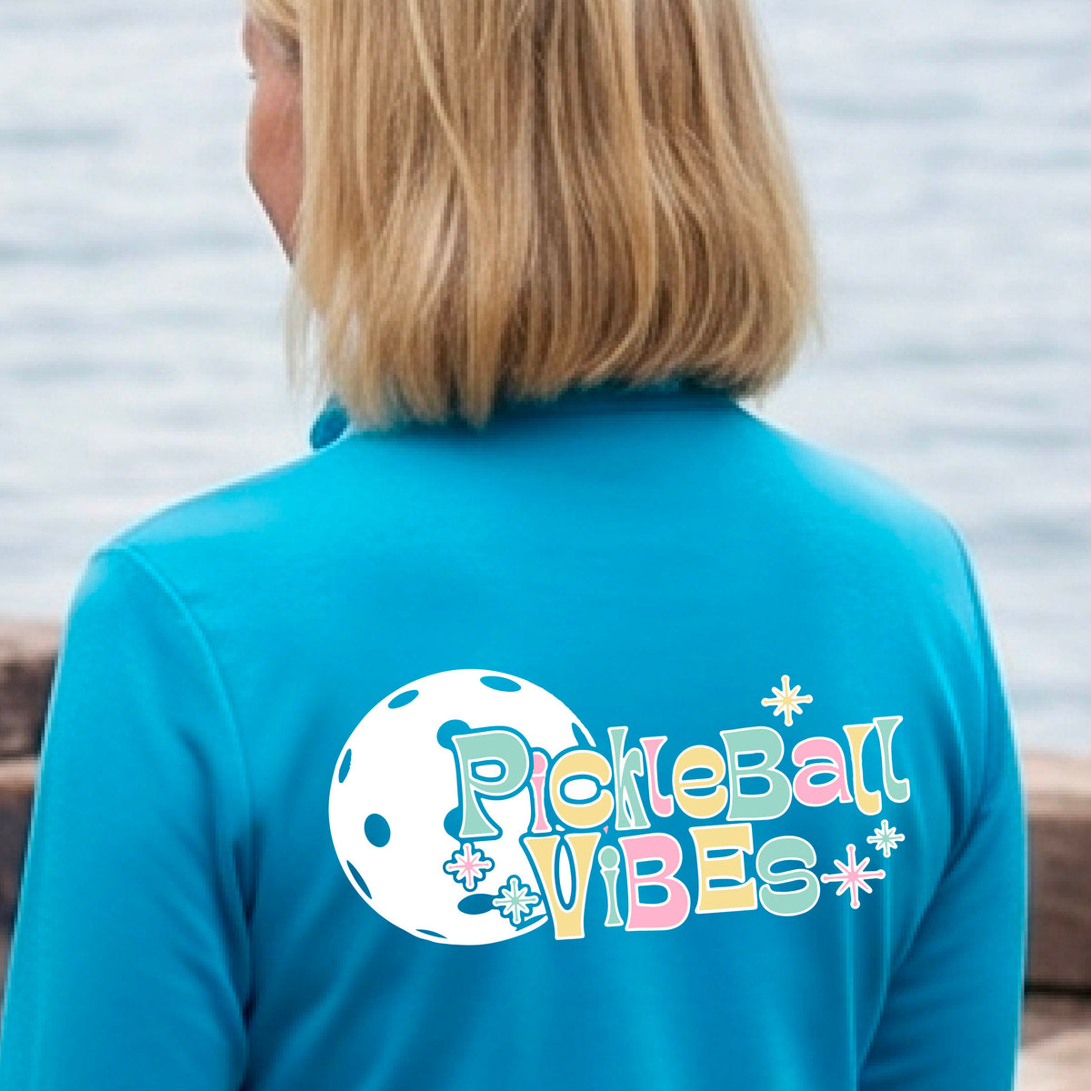 A close-up of a woman, facing backwards on a pickleball court wearing a bright blue quarter-zip shirt, with a colorful retro style font 'Pickleball Vibes' on top of a white pickleball graphic, on the back of the shirt. 