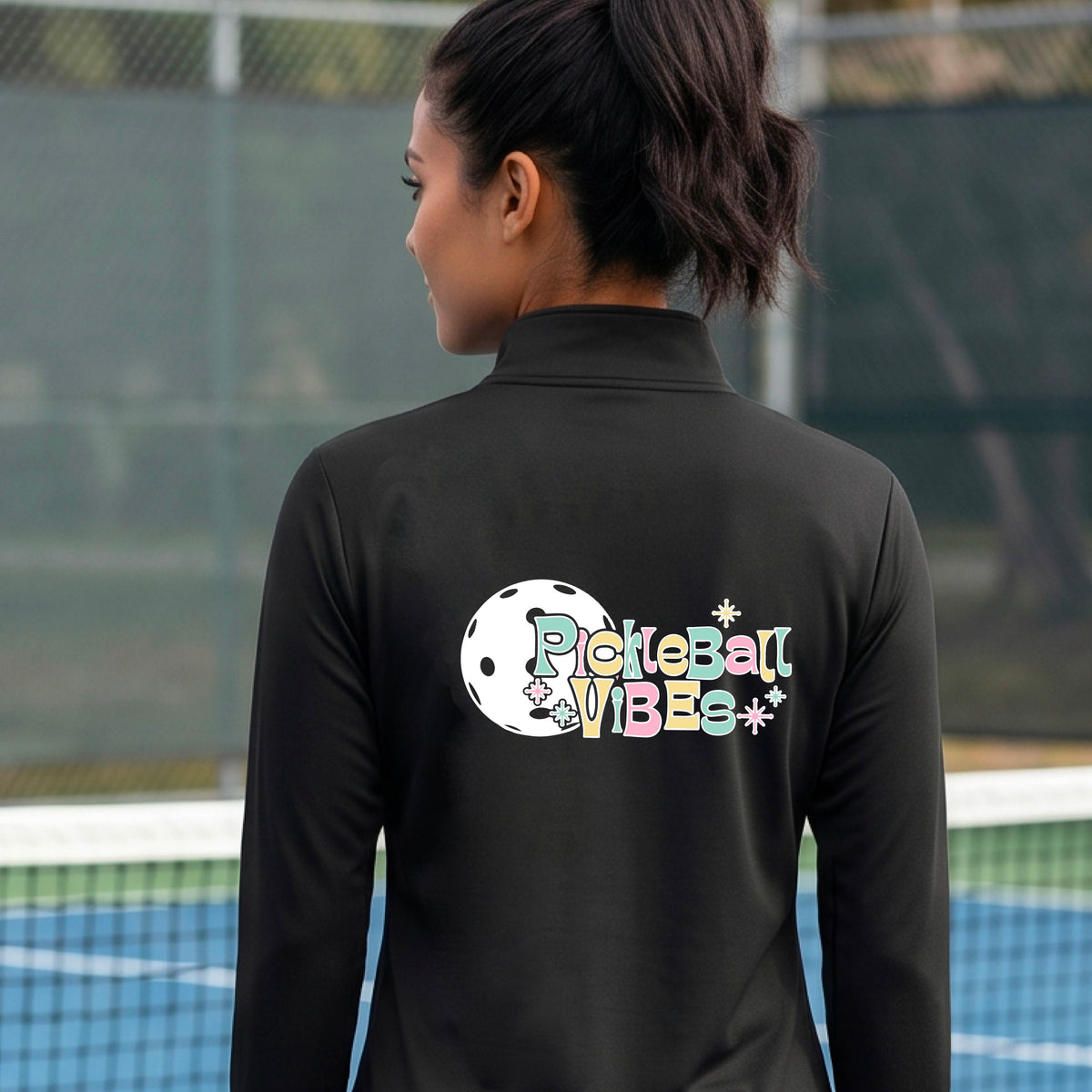 A close-up of a woman, facing backwards on a pickleball court wearing a black quarter-zip shirt, with a colorful retro style font 'Pickleball Vibes' on top of a white pickleball graphic, on the back of the shirt. 