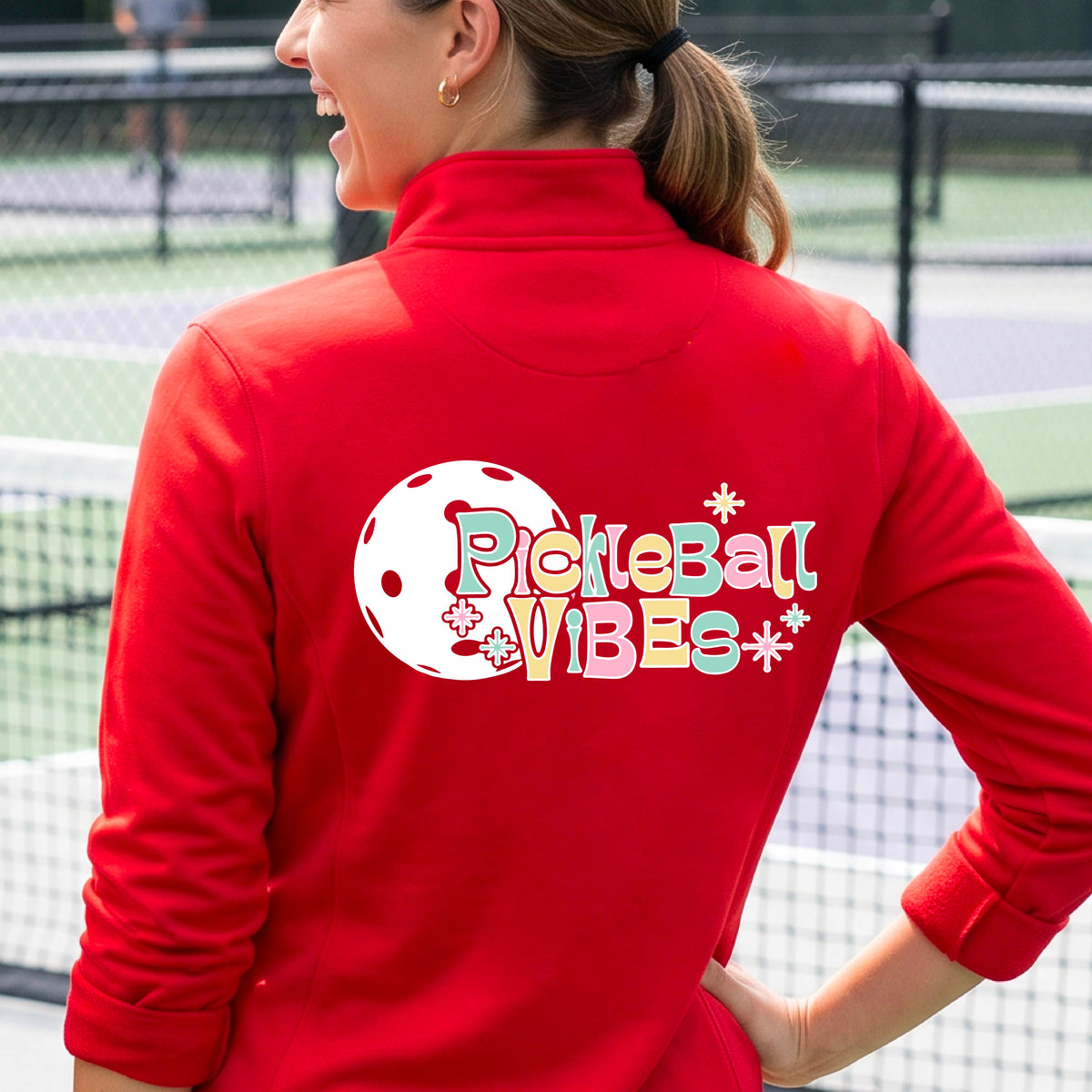 A close-up of a woman, facing backwards on a pickleball court wearing a red quarter-zip shirt, with a colorful retro style font 'Pickleball Vibes' on top of a white pickleball graphic, on the back of the shirt. 