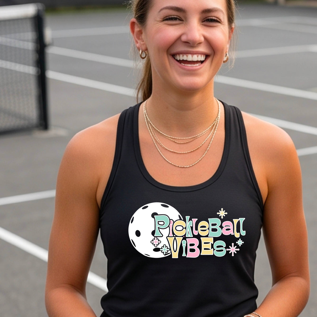A close-up of a woman on a pickleball wearing a black racerbak tank top, with a colorful retro style font 'Pickleball Vibes' on top of a white pickleball graphic, on the front of the shirt.