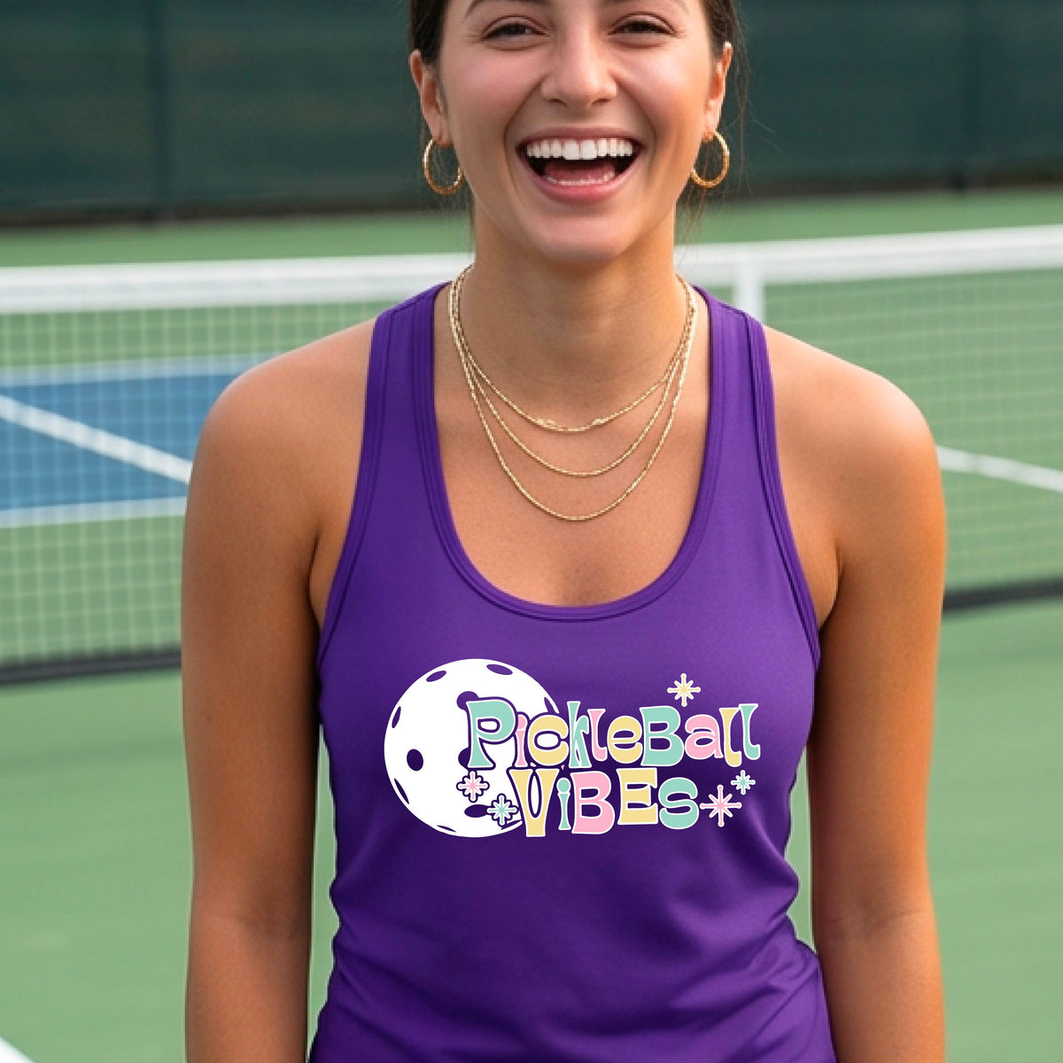 A close-up of a woman on a pickleball wearing a purple racerbak tank top, with a colorful retro style font 'Pickleball Vibes' on top of a white pickleball graphic, on the front of the shirt.