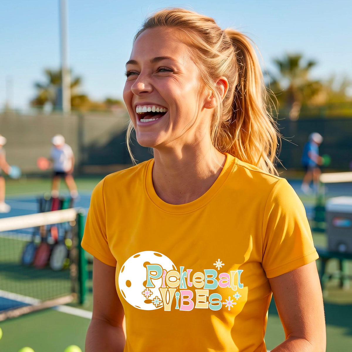 A close-up of a woman on a pickleball wearing a mustard yellow gold short-sleeve t-shirt, with a colorful retro style font 'Pickleball Vibes' on top of a white pickleball graphic, on the front of the shirt.
