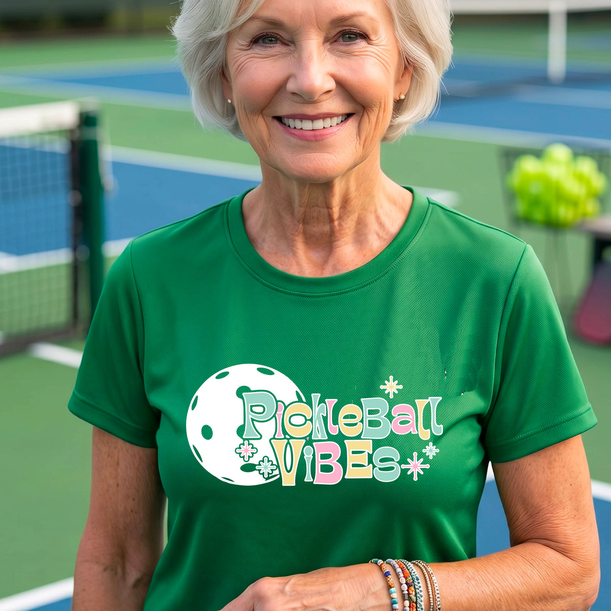 A close-up of a woman on a pickleball wearing a green short-sleeve t-shirt, with a colorful retro style font 'Pickleball Vibes' on top of a white pickleball graphic, on the front of the shirt.