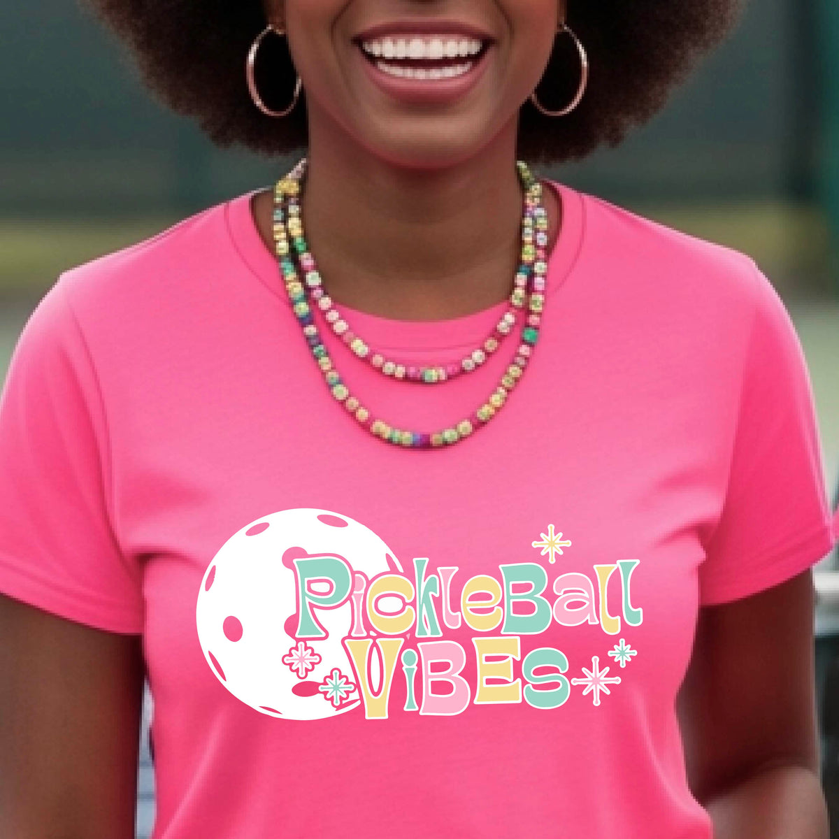 A close-up of a woman on a pickleball wearing a pink short-sleeve t-shirt, with a colorful retro style font 'Pickleball Vibes' on top of a white pickleball graphic, on the front of the shirt.