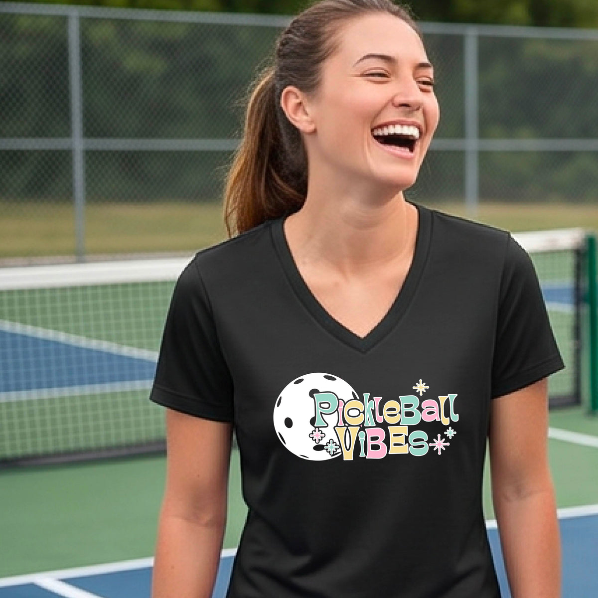 A close-up of a woman on a pickleball wearing a black short-sleeve t-shirt, with a colorful retro style font 'Pickleball Vibes' on top of a white pickleball graphic, on the front of the shirt.