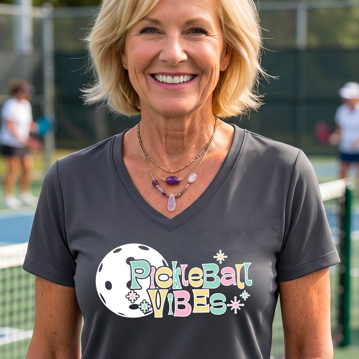 A close-up of a woman on a pickleball wearing a gray short-sleeve t-shirt, with a colorful retro style font 'Pickleball Vibes' on top of a white pickleball graphic, on the front of the shirt.