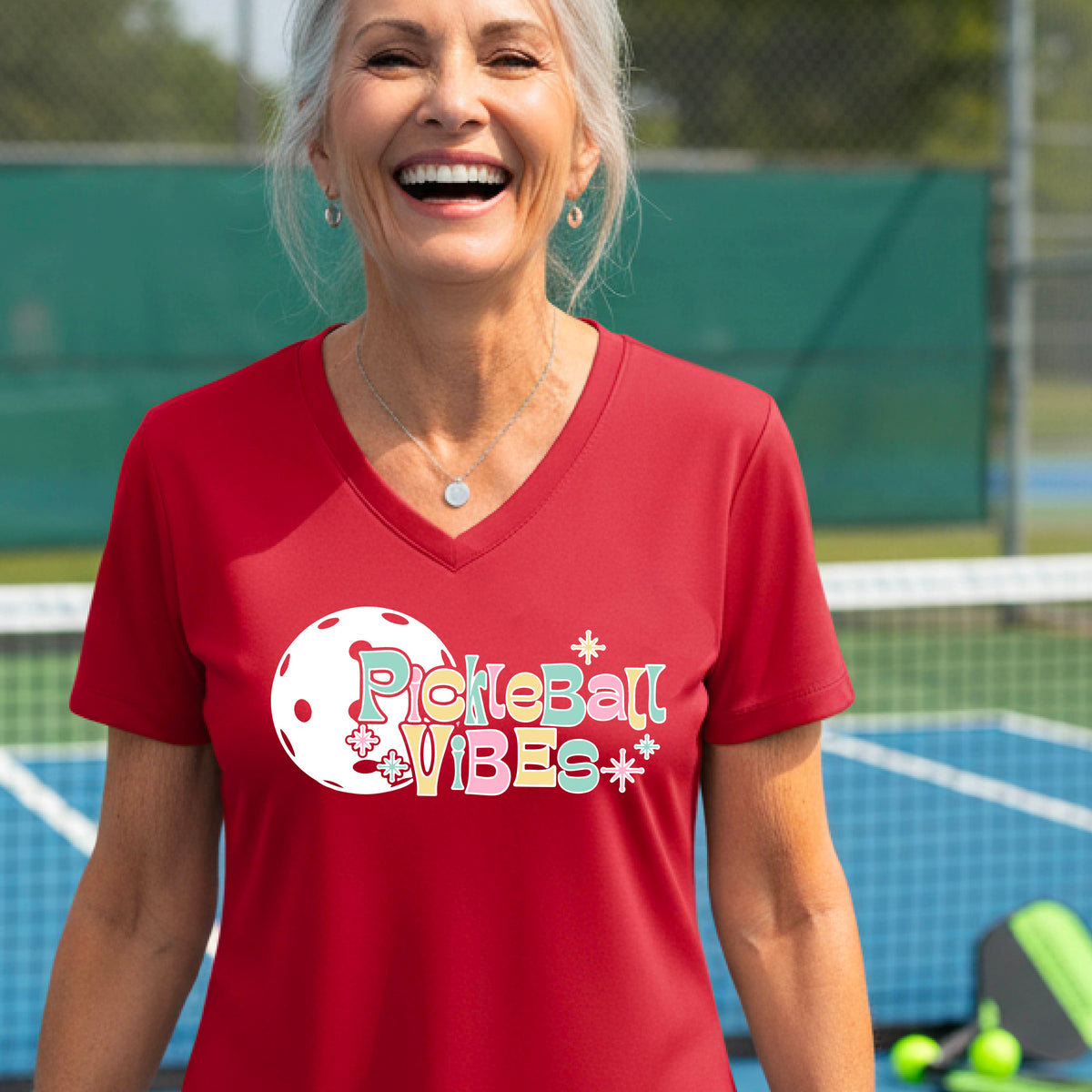 A close-up of a woman on a pickleball wearing a red short-sleeve t-shirt, with a colorful retro style font 'Pickleball Vibes' on top of a white pickleball graphic, on the front of the shirt.