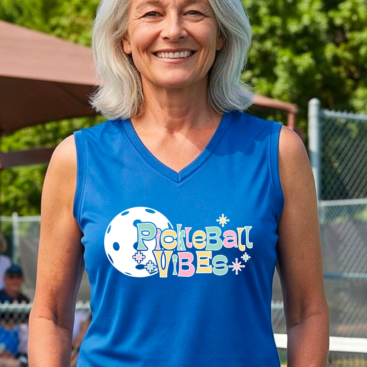 A close-up of a woman on a pickleball wearing a royal blue sleeveless t-shirt, with a colorful retro style font 'Pickleball Vibes' on top of a white pickleball graphic, on the front of the shirt.
