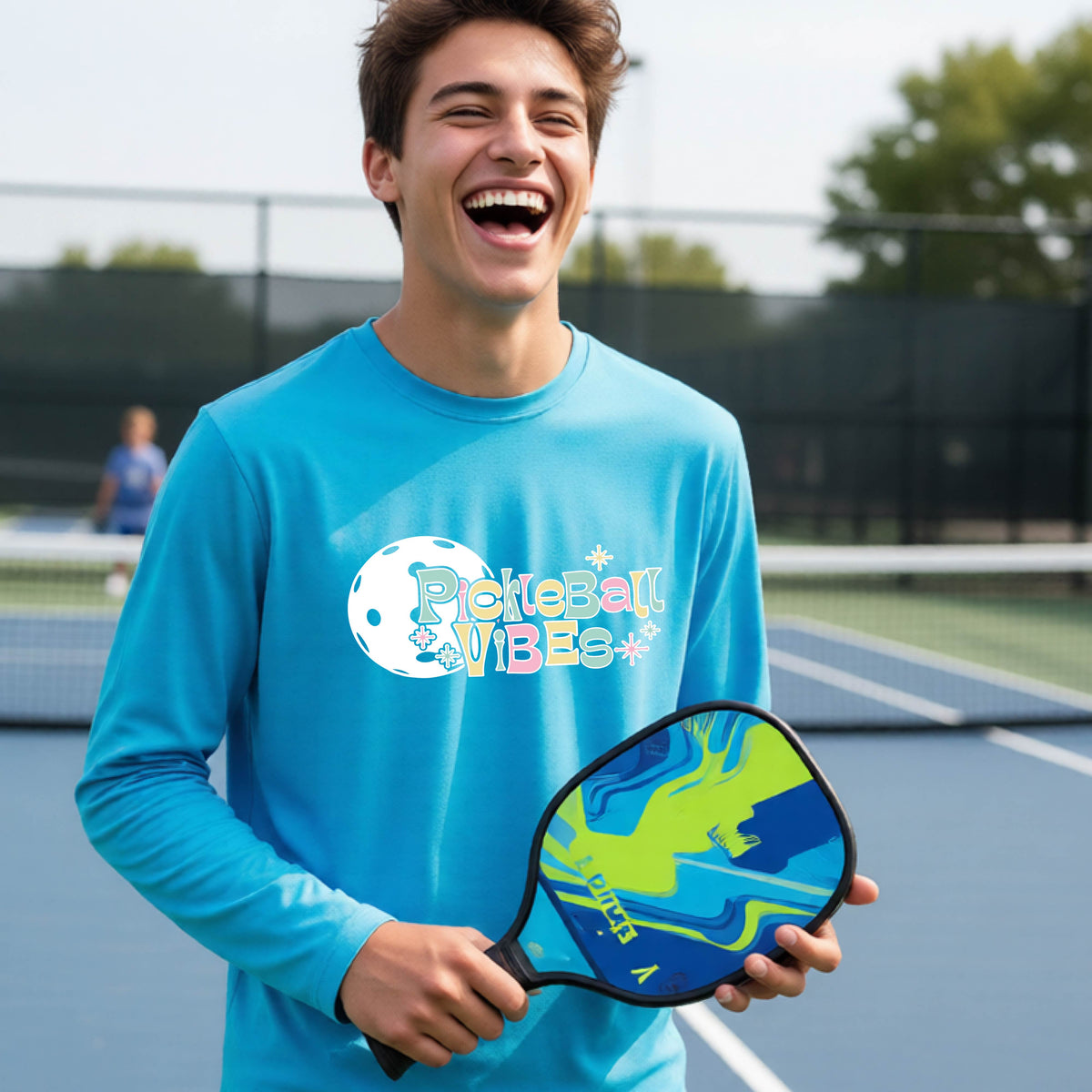 A close-up of a young person on a pickleball wearing a bright blue short-sleeve t-shirt, with a colorful retro style font 'Pickleball Vibes' on top of a white pickleball graphic, on the front of the shirt.