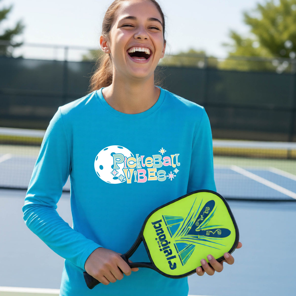 A close-up of a young person on a pickleball wearing a bright blue short-sleeve t-shirt, with a colorful retro style font 'Pickleball Vibes' on top of a white pickleball graphic, on the front of the shirt.