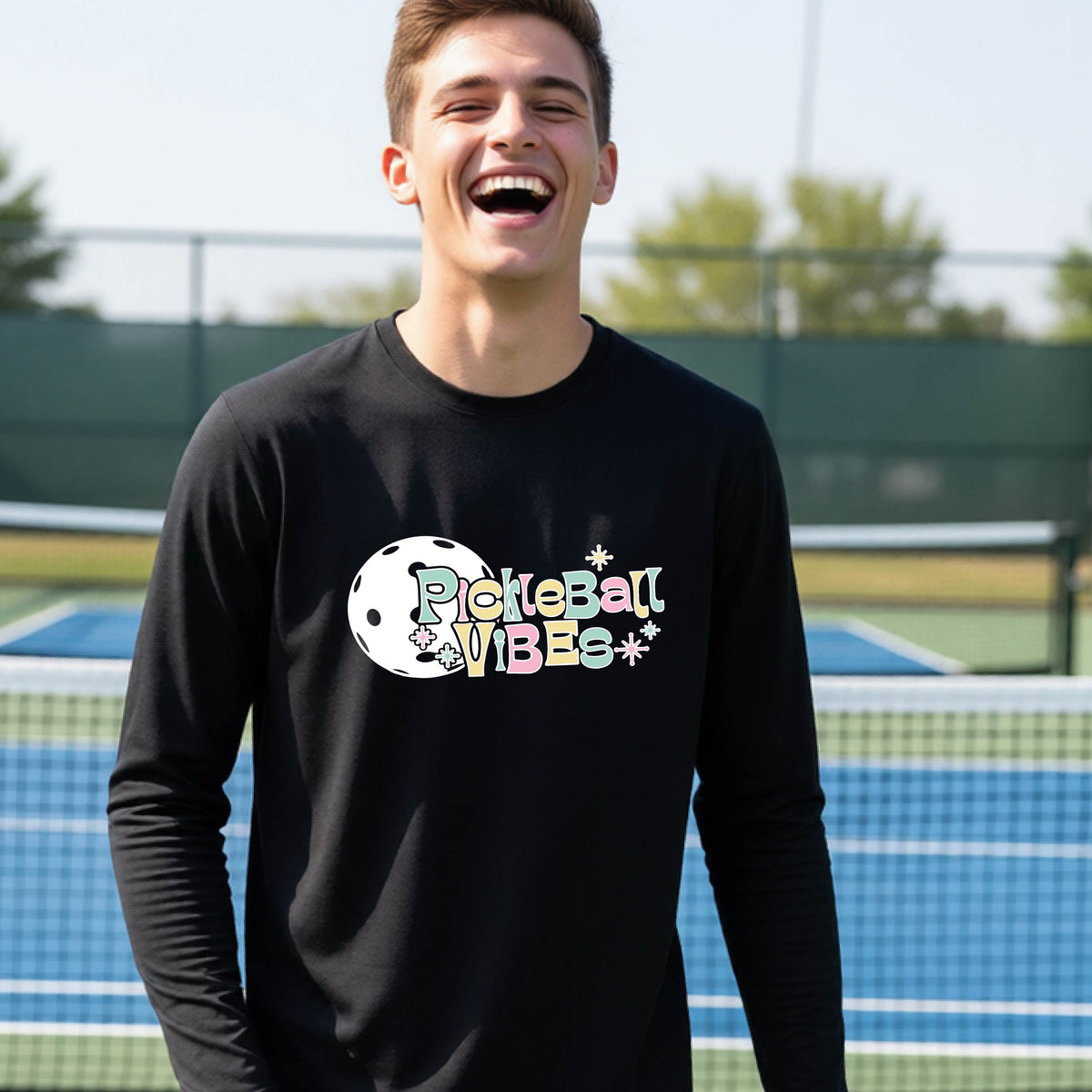 A close-up of a young person on a pickleball wearing a black short-sleeve t-shirt, with a colorful retro style font 'Pickleball Vibes' on top of a white pickleball graphic, on the front of the shirt.