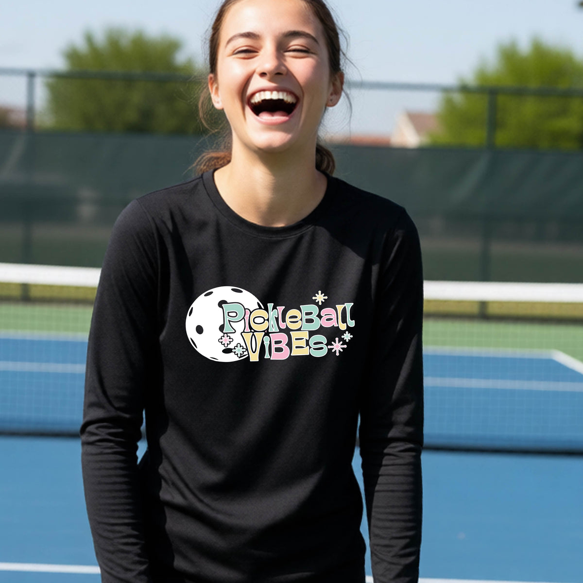 A close-up of a young person on a pickleball wearing a black short-sleeve t-shirt, with a colorful retro style font 'Pickleball Vibes' on top of a white pickleball graphic, on the front of the shirt.