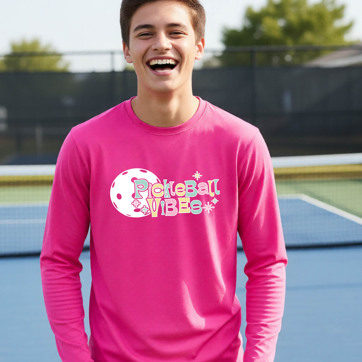 A close-up of a young person on a pickleball wearing a pink short-sleeve t-shirt, with a colorful retro style font 'Pickleball Vibes' on top of a white pickleball graphic, on the front of the shirt.