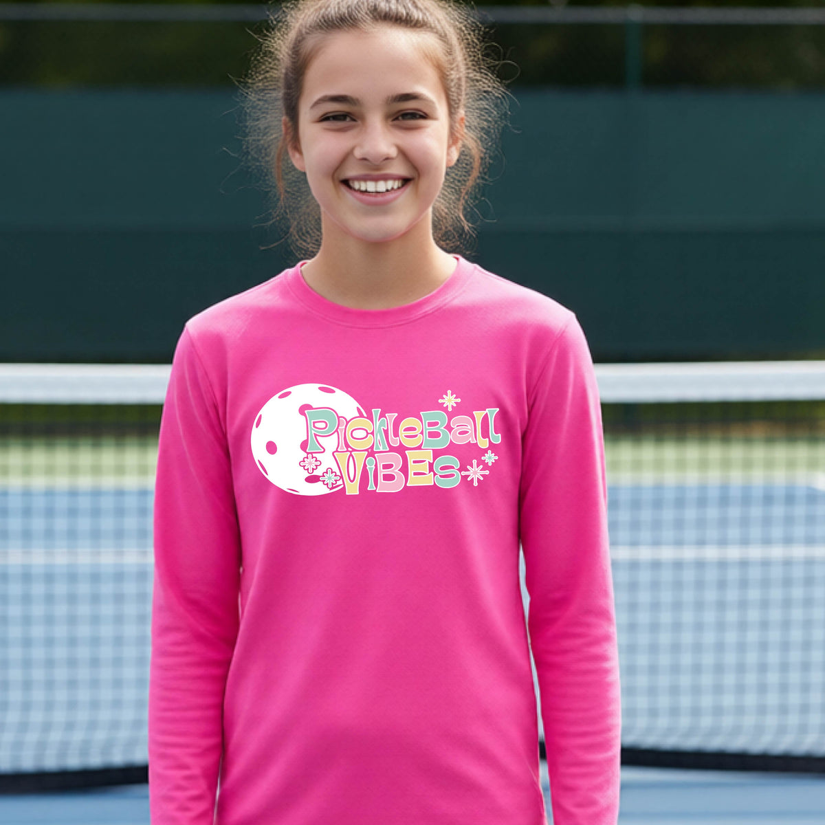 A close-up of a young person on a pickleball wearing a pink short-sleeve t-shirt, with a colorful retro style font 'Pickleball Vibes' on top of a white pickleball graphic, on the front of the shirt.