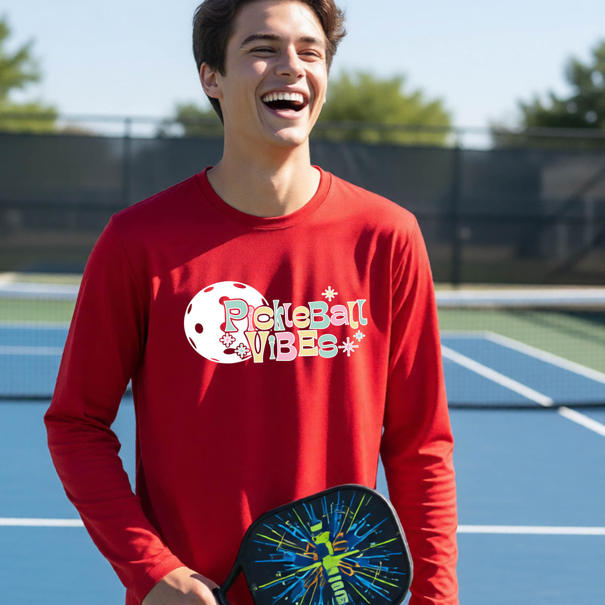 A close-up of a young person on a pickleball wearing a red short-sleeve t-shirt, with a colorful retro style font 'Pickleball Vibes' on top of a white pickleball graphic, on the front of the shirt.