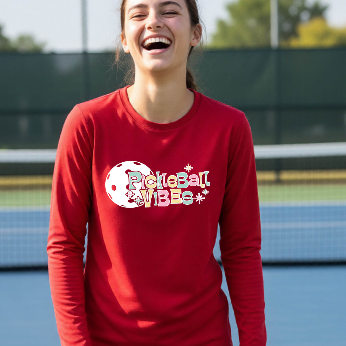 A close-up of a young person on a pickleball wearing a red short-sleeve t-shirt, with a colorful retro style font 'Pickleball Vibes' on top of a white pickleball graphic, on the front of the shirt.