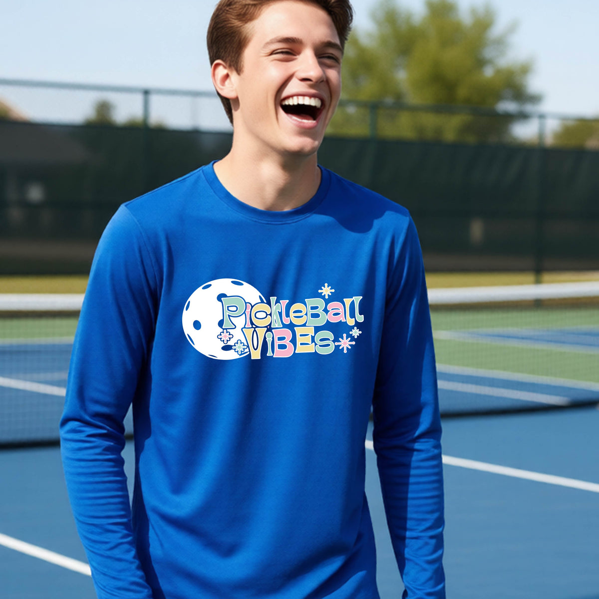 A close-up of a young person on a pickleball wearing a royal blue short-sleeve t-shirt, with a colorful retro style font 'Pickleball Vibes' on top of a white pickleball graphic, on the front of the shirt.