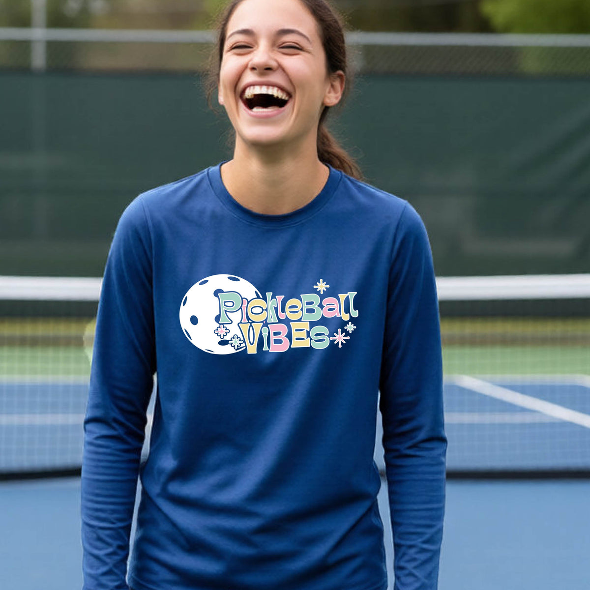A close-up of a young person on a pickleball wearing a royal blue short-sleeve t-shirt, with a colorful retro style font 'Pickleball Vibes' on top of a white pickleball graphic, on the front of the shirt.