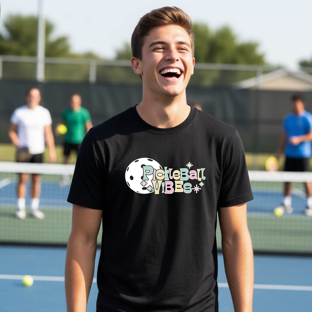 A close-up of a young person on a pickleball wearing a black short-sleeve t-shirt, with a colorful retro style font 'Pickleball Vibes' on top of a white pickleball graphic, on the front of the shirt.