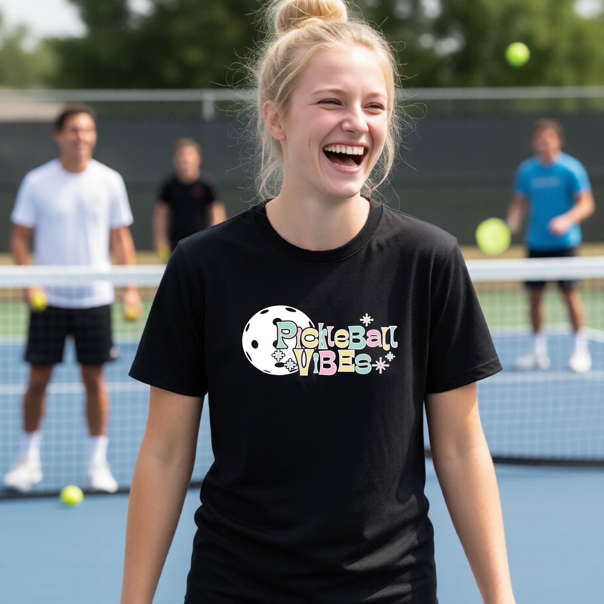 A close-up of a young person on a pickleball wearing a black short-sleeve t-shirt, with a colorful retro style font 'Pickleball Vibes' on top of a white pickleball graphic, on the front of the shirt.