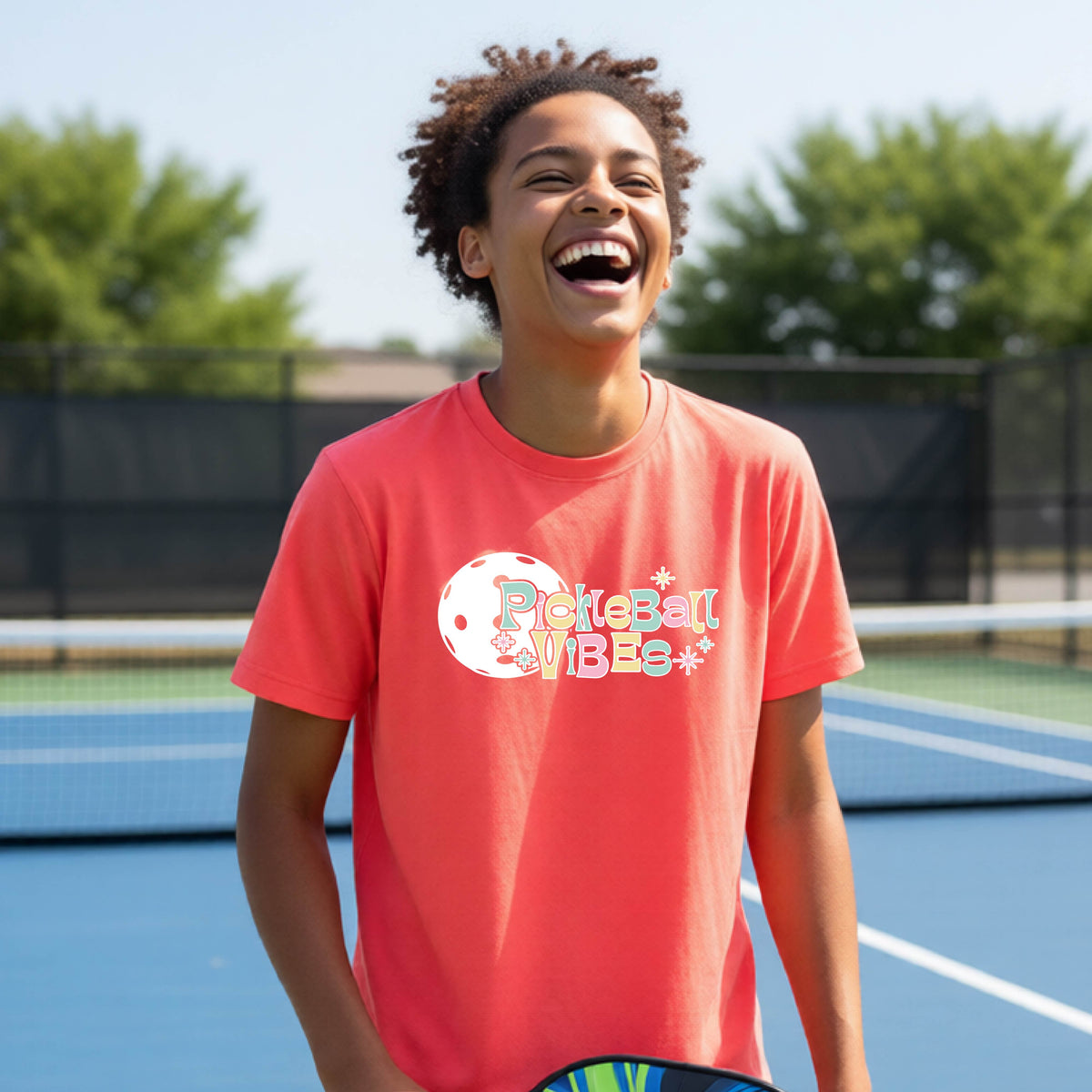 A close-up of a young person on a pickleball wearing a coral pink short-sleeve t-shirt, with a colorful retro style font 'Pickleball Vibes' on top of a white pickleball graphic, on the front of the shirt.