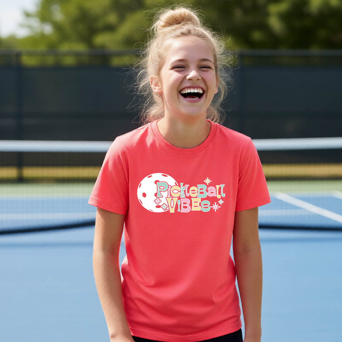 A close-up of a young person on a pickleball wearing a coral pink short-sleeve t-shirt, with a colorful retro style font 'Pickleball Vibes' on top of a white pickleball graphic, on the front of the shirt.