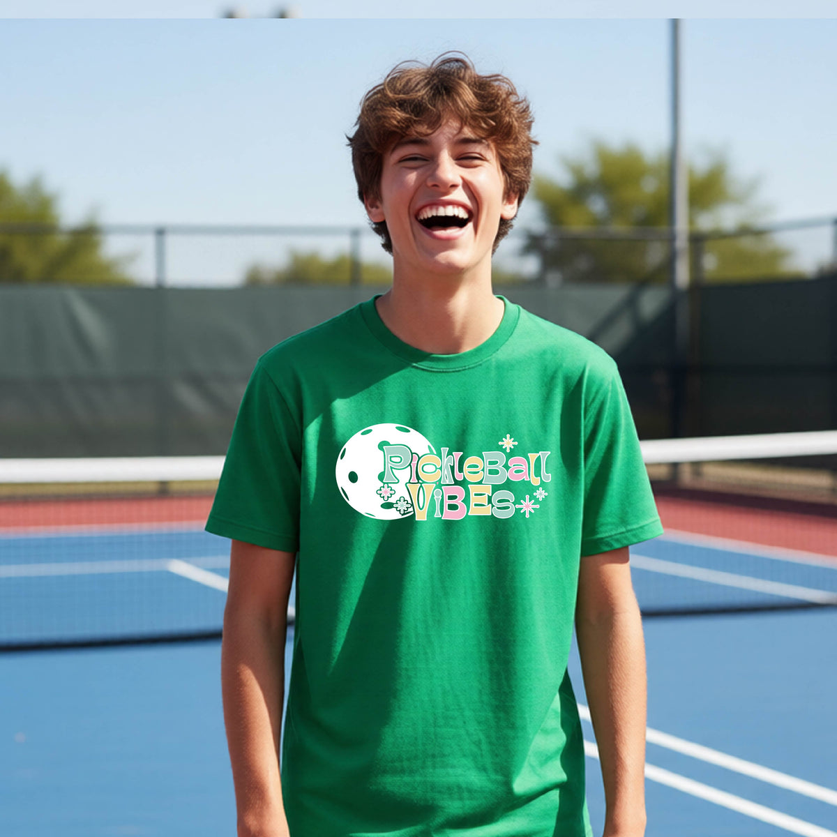 A close-up of a young person on a pickleball wearing a green short-sleeve t-shirt, with a colorful retro style font 'Pickleball Vibes' on top of a white pickleball graphic, on the front of the shirt.