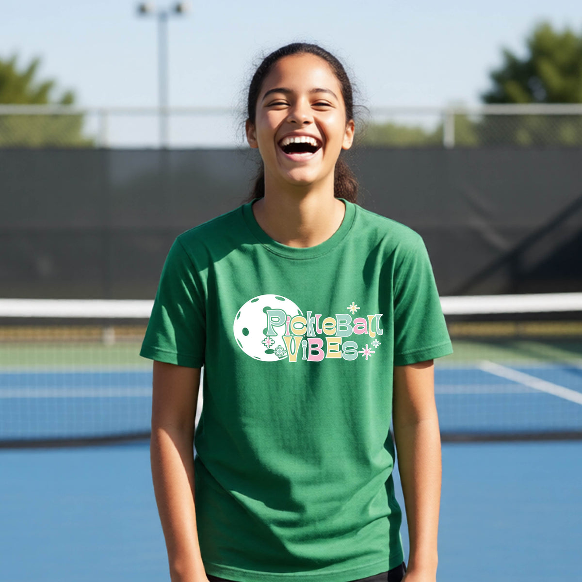 A close-up of a young person on a pickleball wearing a green short-sleeve t-shirt, with a colorful retro style font 'Pickleball Vibes' on top of a white pickleball graphic, on the front of the shirt.