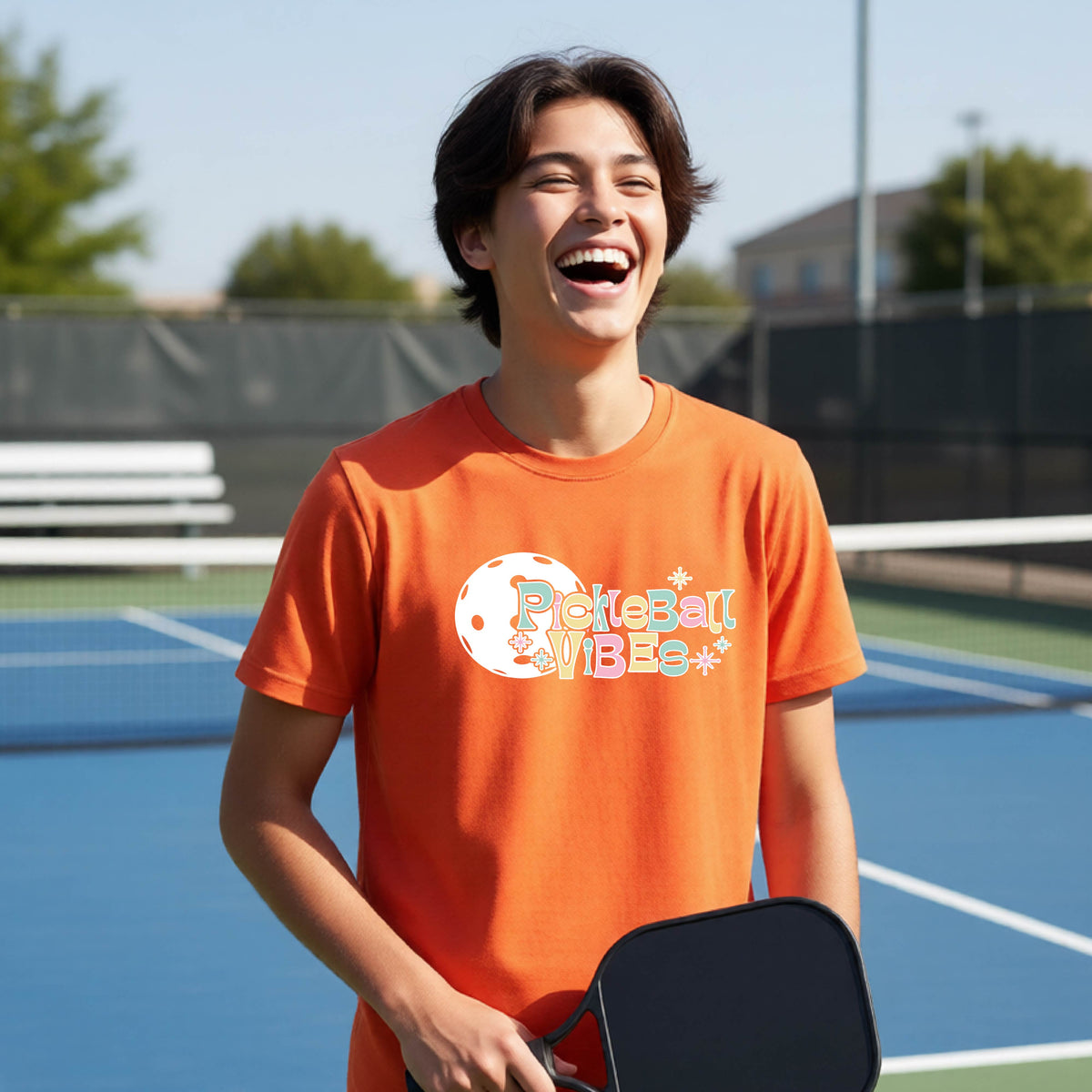 A close-up of a young person on a pickleball wearing an orange short-sleeve t-shirt, with a colorful retro style font 'Pickleball Vibes' on top of a white pickleball graphic, on the front of the shirt.