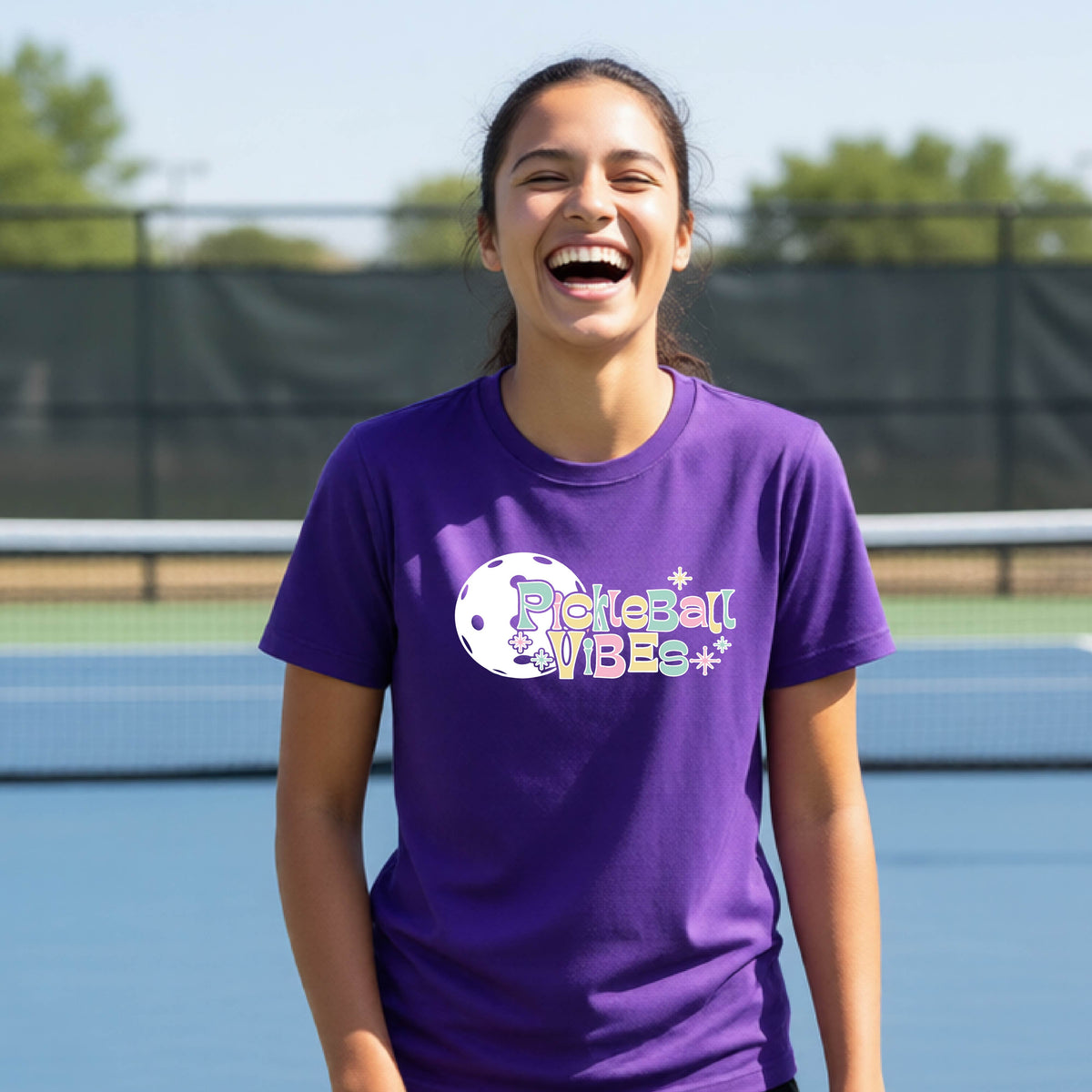 A close-up of a young person on a pickleball wearing a purple short-sleeve t-shirt, with a colorful retro style font 'Pickleball Vibes' on top of a white pickleball graphic, on the front of the shirt.