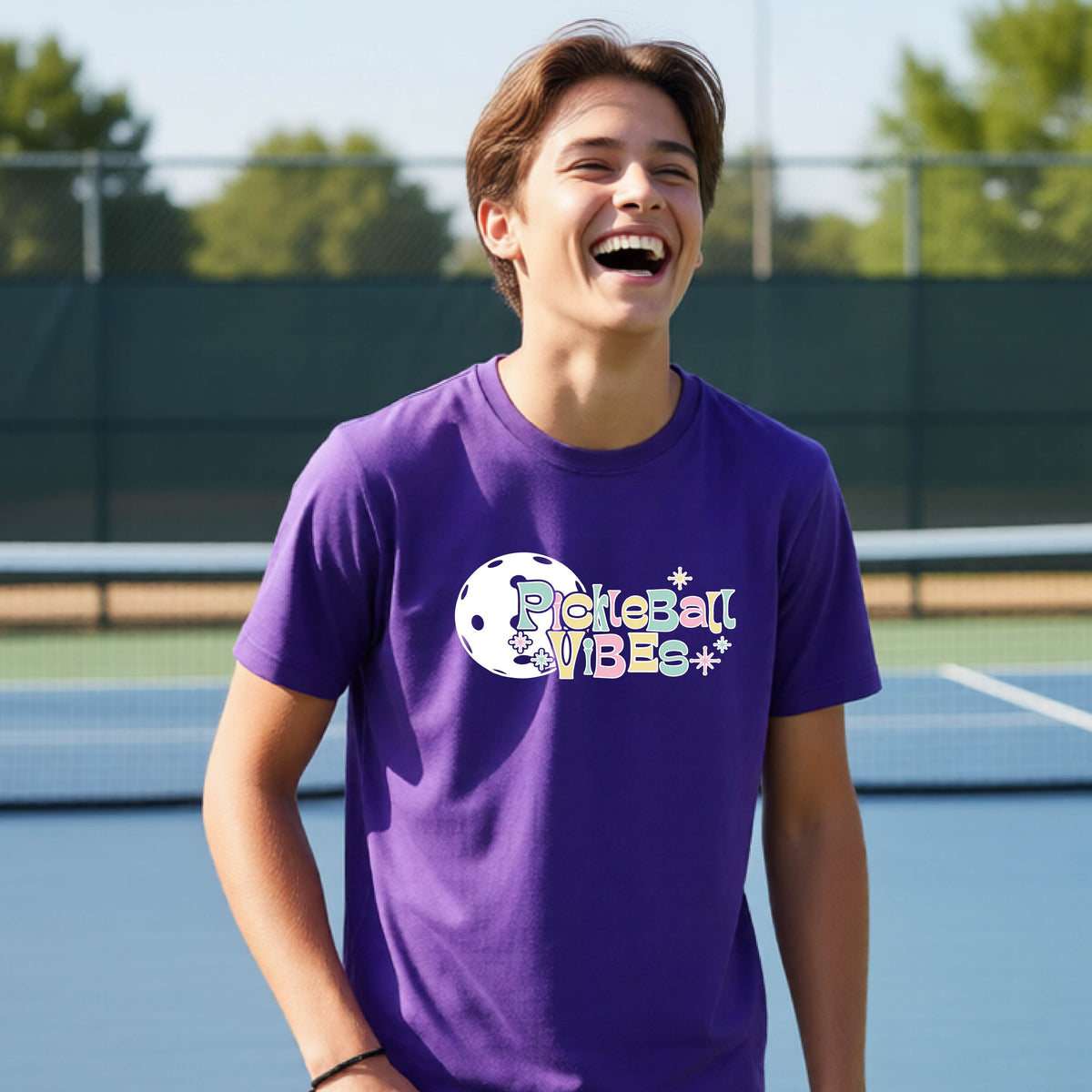 A close-up of a young person on a pickleball wearing a purple short-sleeve t-shirt, with a colorful retro style font 'Pickleball Vibes' on top of a white pickleball graphic, on the front of the shirt.