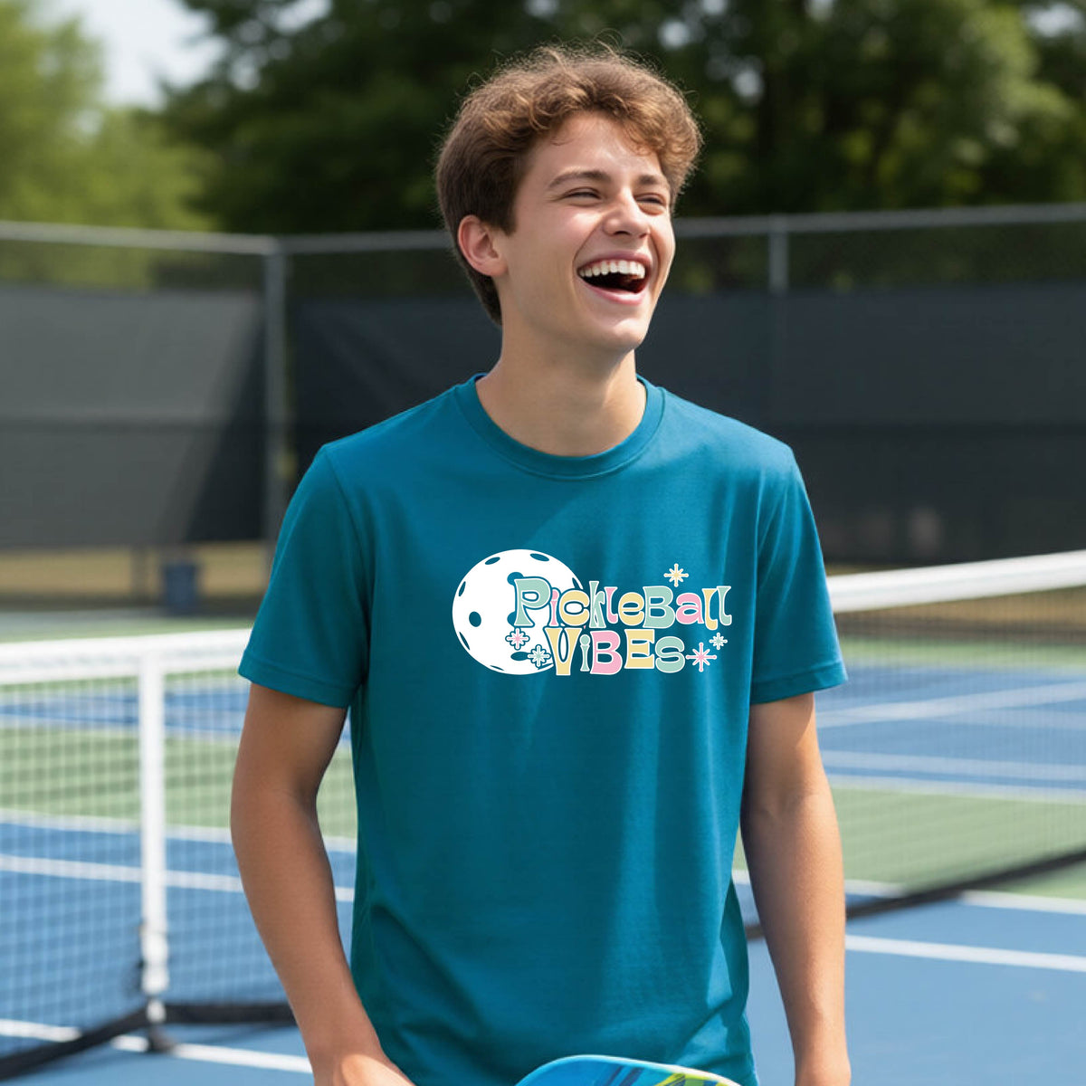A close-up of a young person on a pickleball wearing a teal short-sleeve t-shirt, with a colorful retro style font 'Pickleball Vibes' on top of a white pickleball graphic, on the front of the shirt.