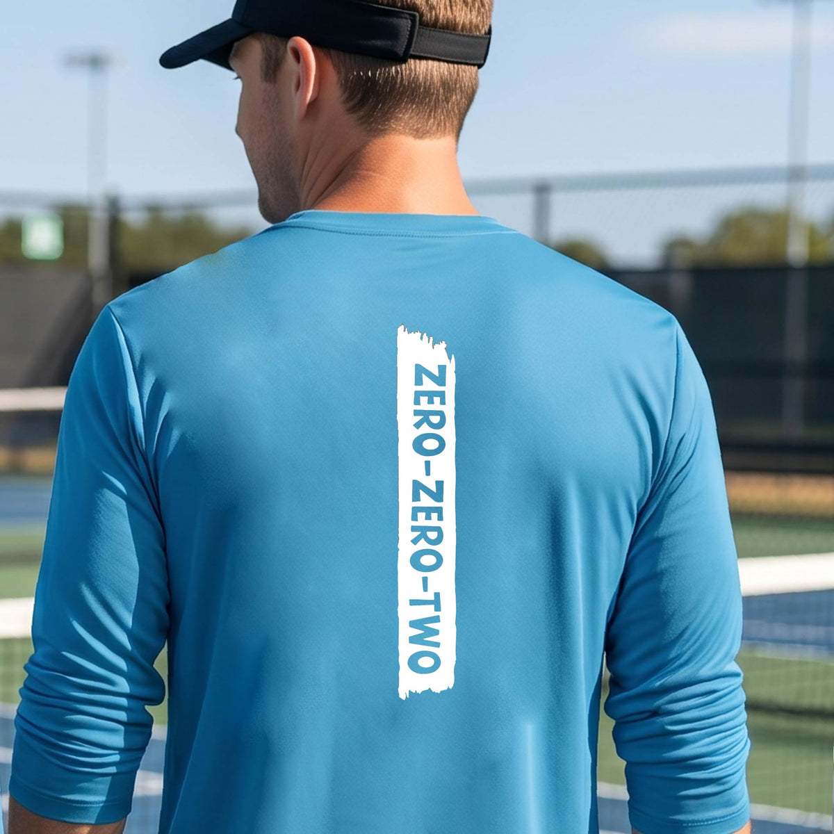 A close-up of a man on a pickleball wearing a bright blue long-sleeve t-shirt, with a white graphic of artistic banner with the cutout words of ‘ZERO ZERO TWO’, on the back of the shirt. 