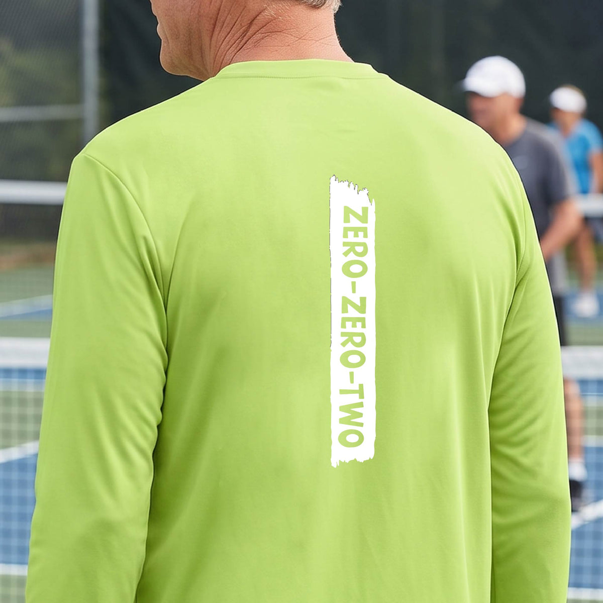 A close-up of a man on a pickleball wearing a lime green long-sleeve t-shirt, with a white graphic of artistic banner with the cutout words of ‘ZERO ZERO TWO’, on the back of the shirt. 