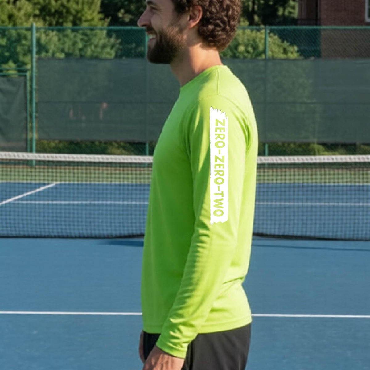 A close-up of a man on a pickleball wearing a lime green long-sleeve t-shirt, with a white graphic of artistic banner with the cutout words of ‘ZERO ZERO TWO’, on the sleeve of the shirt. 