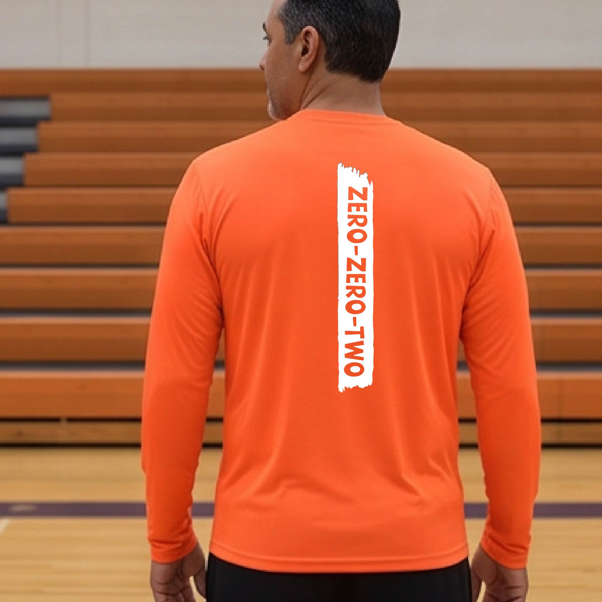 A close-up of a man on a pickleball wearing an orange long-sleeve t-shirt, with a white graphic of artistic banner with the cutout words of ‘ZERO ZERO TWO’, on the back of the shirt. 