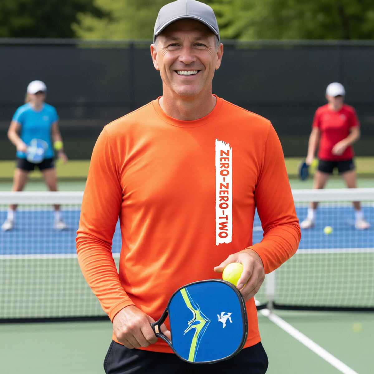 A close-up of a man on a pickleball wearing an orange long-sleeve t-shirt, with a white graphic of artistic banner with the cutout words of ‘ZERO ZERO TWO’, on the front of the shirt. 