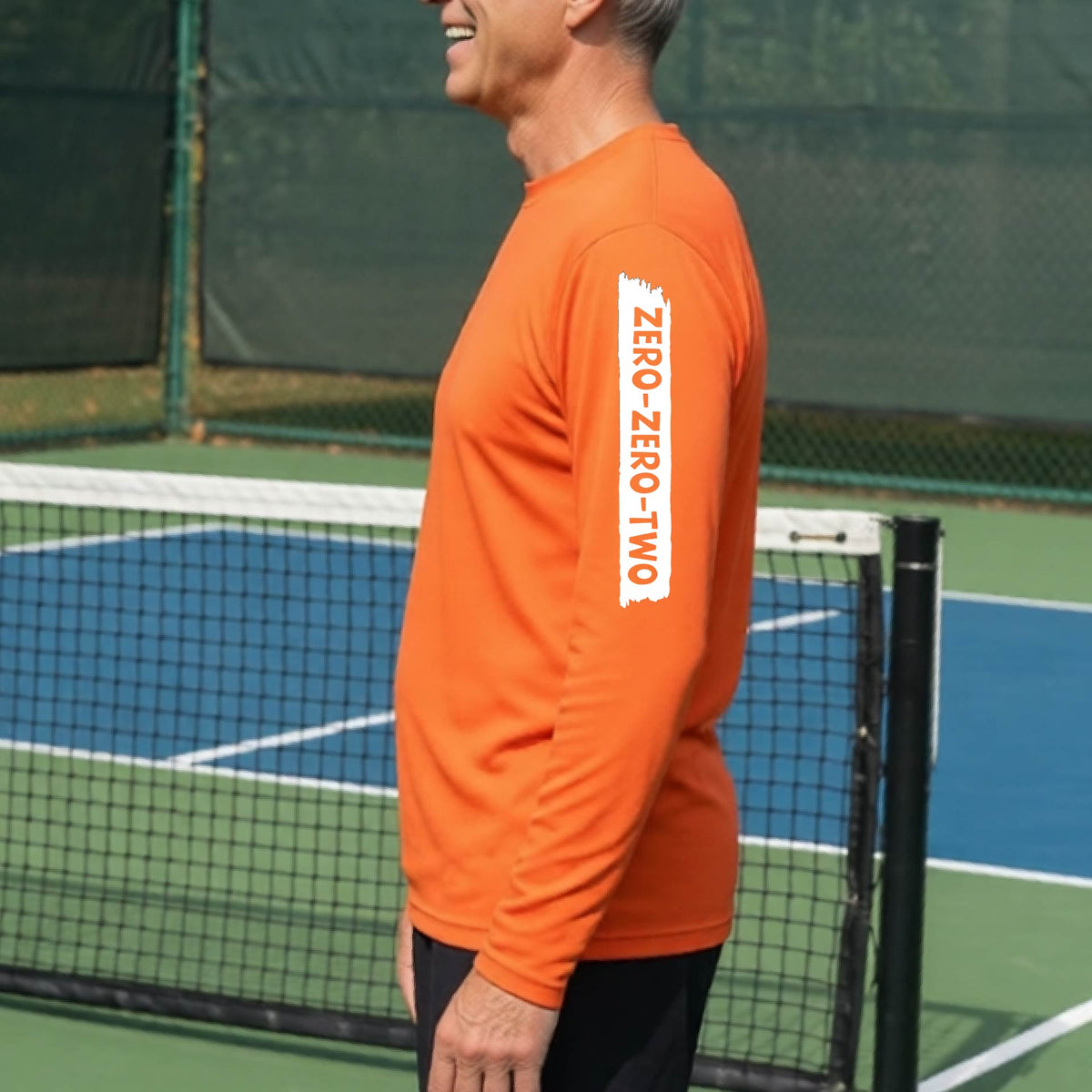 A close-up of a man on a pickleball wearing an orange long-sleeve t-shirt, with a white graphic of artistic banner with the cutout words of ‘ZERO ZERO TWO’, on the sleeve of the shirt. 