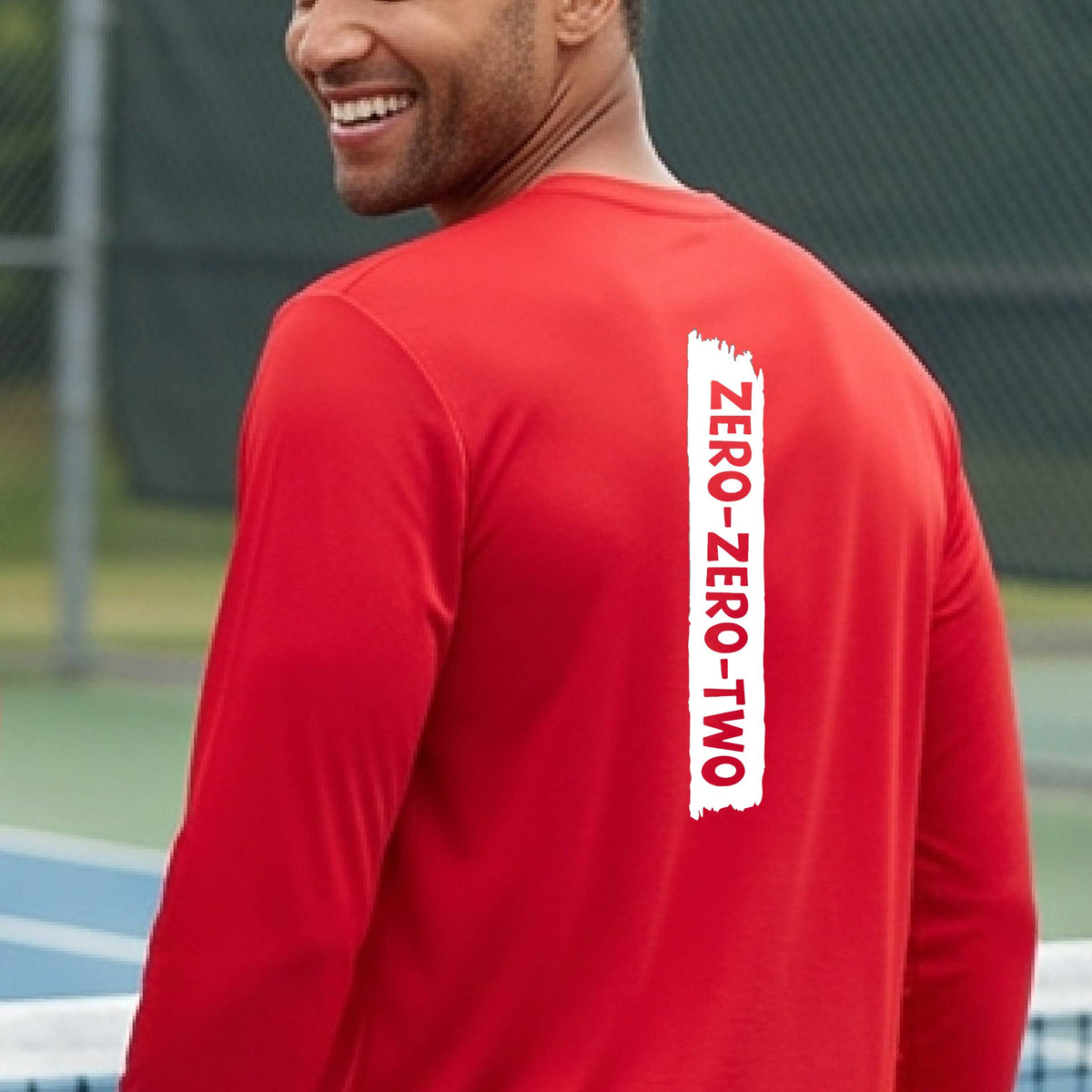 A close-up of a man on a pickleball wearing a red long-sleeve t-shirt, with a white graphic of artistic banner with the cutout words of ‘ZERO ZERO TWO’, on the back of the shirt. 