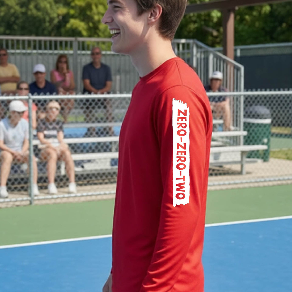 A close-up of a man on a pickleball wearing a red long-sleeve t-shirt, with a white graphic of artistic banner with the cutout words of ‘ZERO ZERO TWO’, on the sleeve of the shirt. 