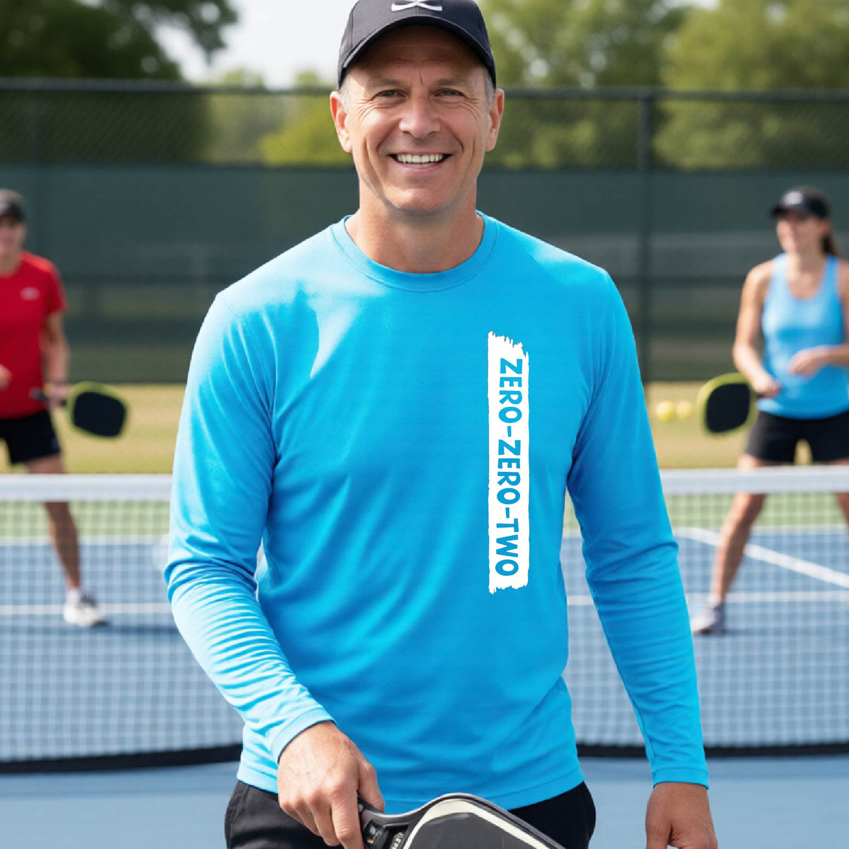 A close-up of a man on a pickleball wearing a bright blue long-sleeve t-shirt, with a white graphic of artistic banner with the cutout words of ‘ZERO ZERO TWO’, on the front of the shirt. 