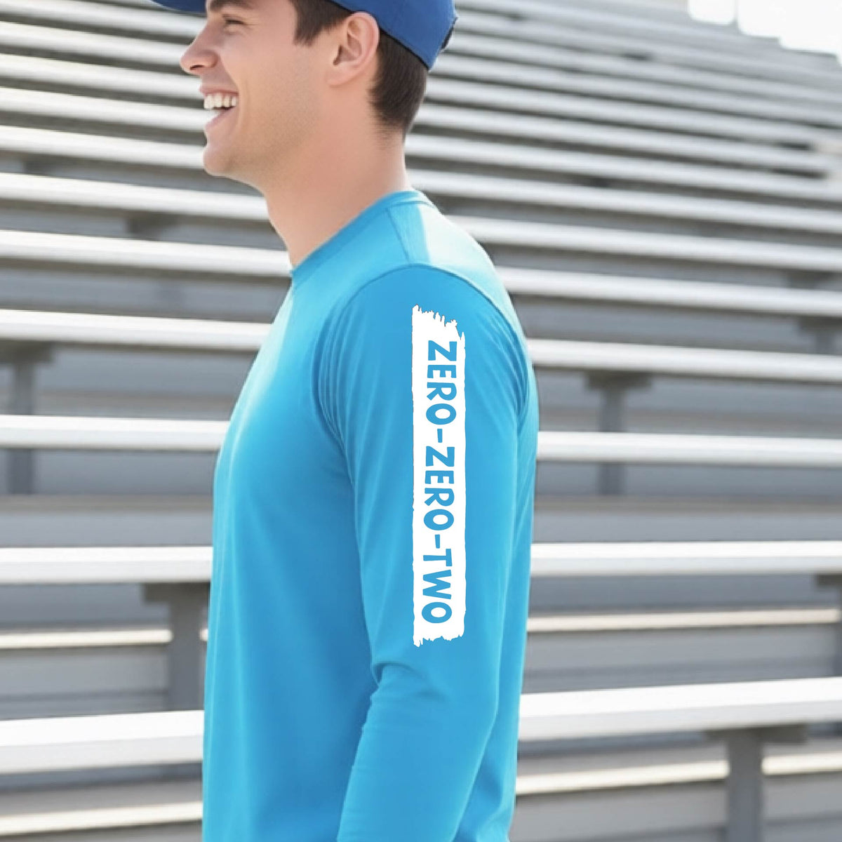 A close-up of a man wearing a bright blue long-sleeve t-shirt, with a white graphic of artistic banner with the cutout words of ‘ZERO ZERO TWO’, on the sleeve of the shirt. 
