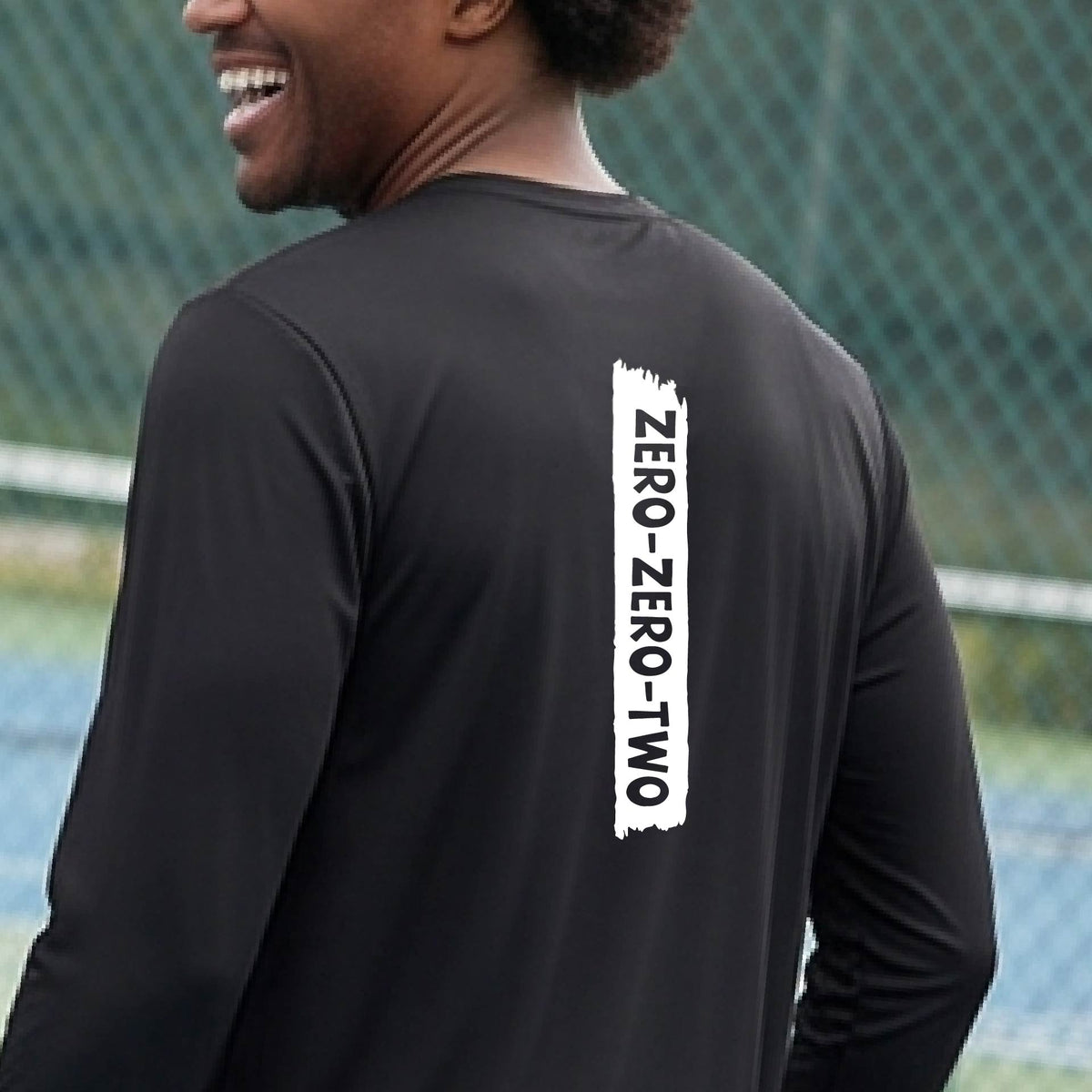 A close-up of a man on a pickleball wearing a black long-sleeve t-shirt, with a white graphic of artistic banner with the cutout words of ‘ZERO ZERO TWO’, on the back of the shirt. 
